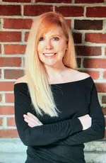 Smiling blonde woman in black off-shoulder top standing with arms crossed against a brick wall background