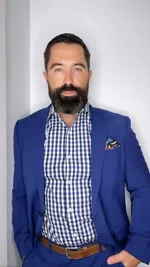 Man with beard wearing blue suit and checkered shirt standing against a plain wall with pocket square