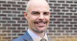 Smiling bald man with a goatee wearing a blue suit and tie standing in front of a brick wall outdoors