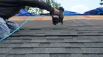 A person using a nail gun to install asphalt shingles on a roof under a clear blue sky.