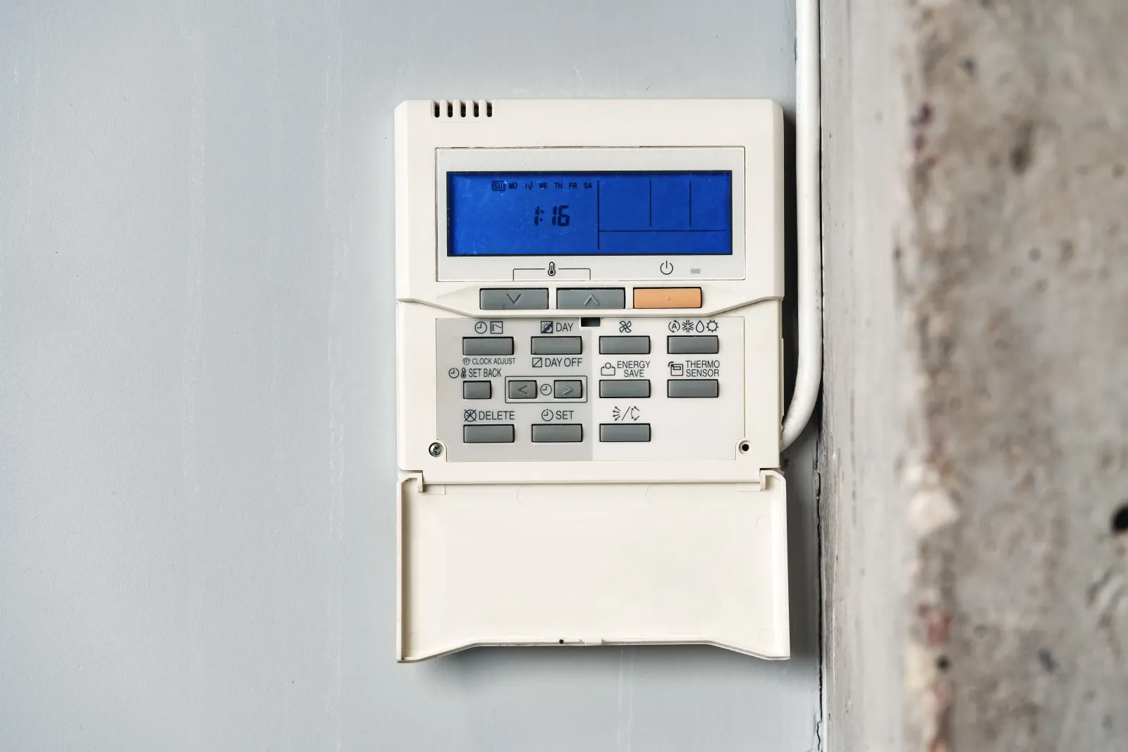 Wall-mounted digital thermostat with blue display screen and multiple buttons on a gray wall indoors.