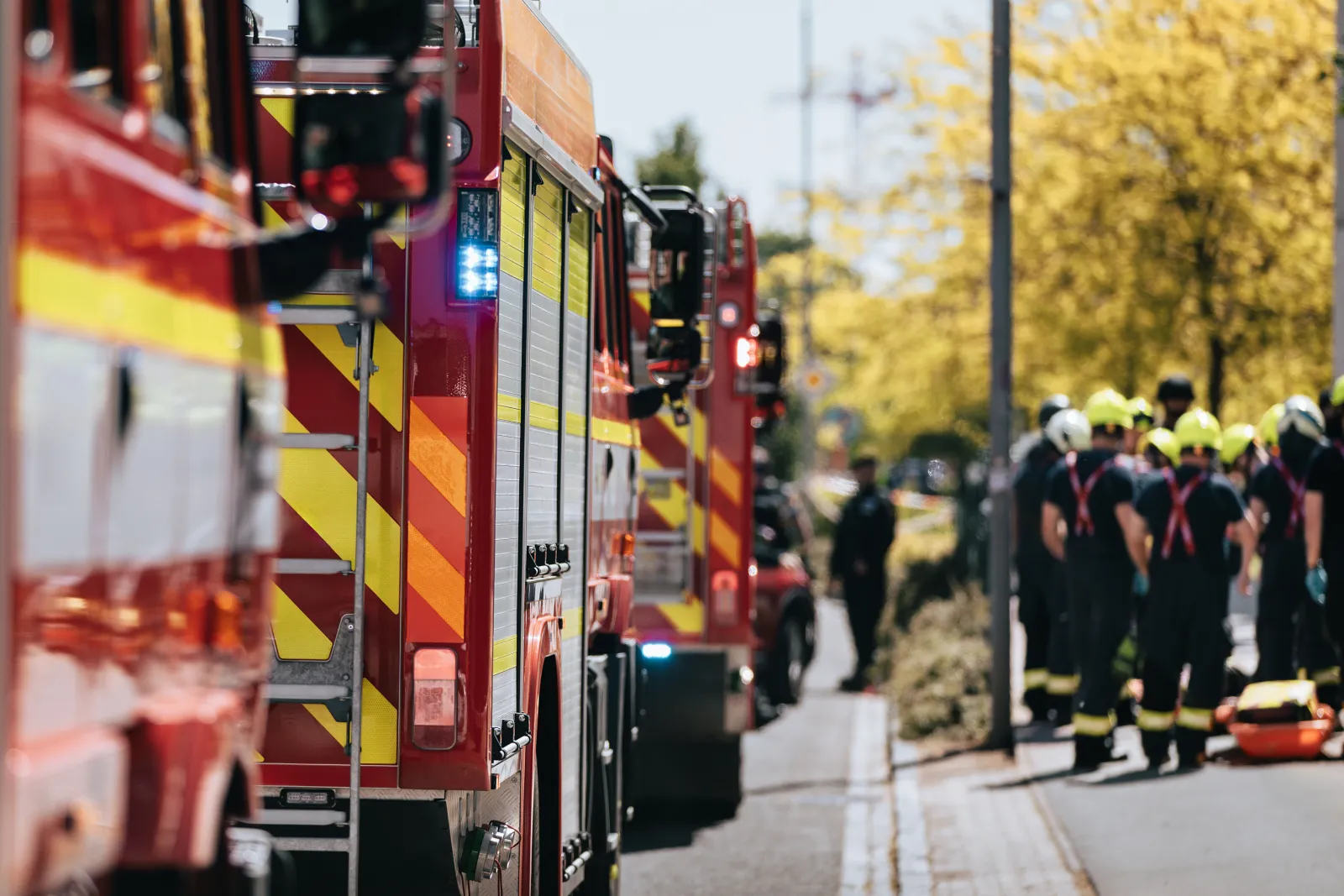 Line of red fire trucks with flashing lights parked on street as firefighters gather in background on sunny day
