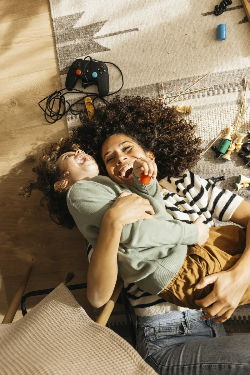 Mother and child laughing and hugging on the floor near game controllers and chess pieces scattered on a rug.