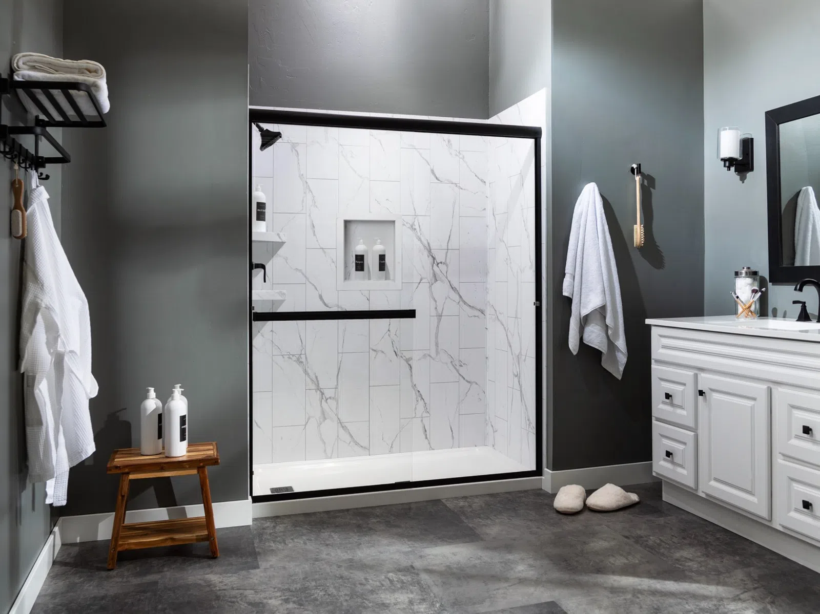 Modern bathroom with dark gray walls, white marble shower, white vanity, and wooden stool with toiletries.