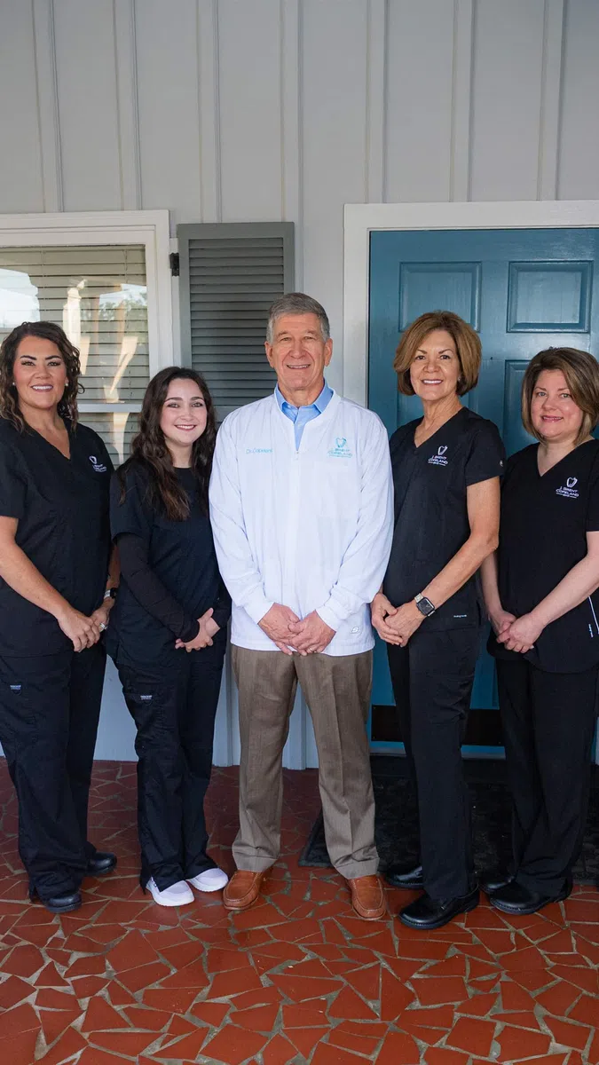 Dental team of five professionals standing on a tiled porch in front of blue door smiling at camera.