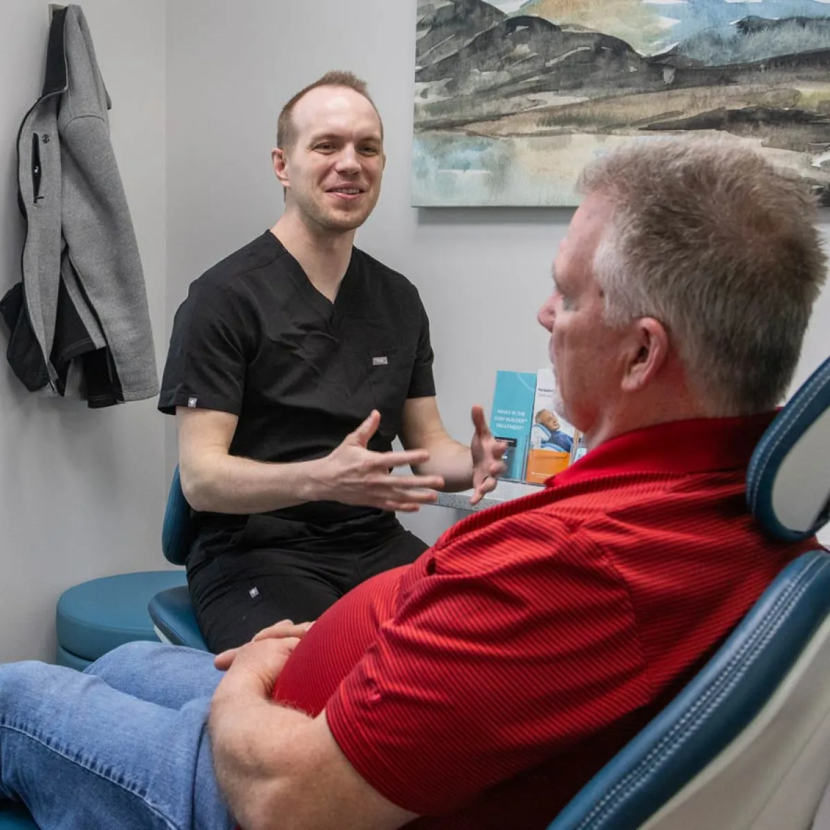 Healthcare professional in black scrubs consulting a male patient in a red shirt in a medical office setting.