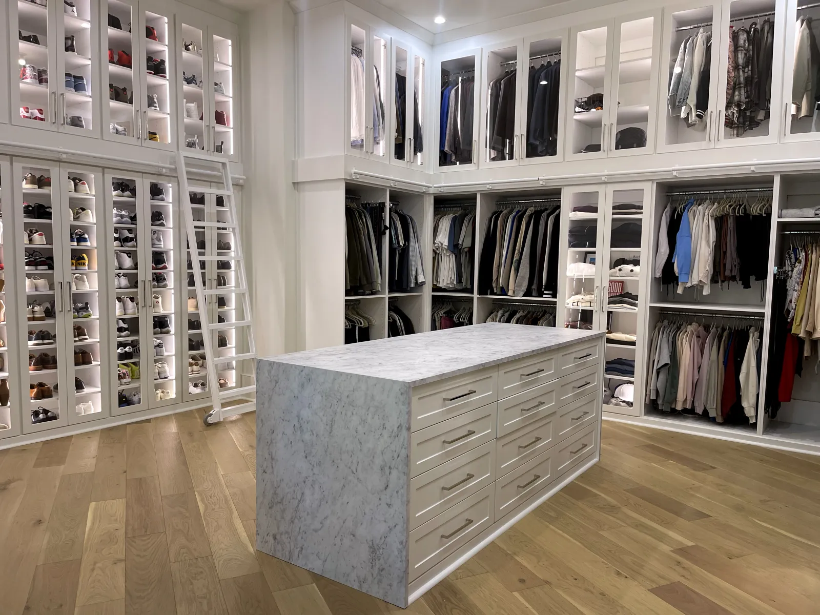 Spacious custom walk in closet, dream closet with marble island with drawers, shoes and sneakers custom closet with glass doors, white shelves with lighting, hanging clothes and white ladder