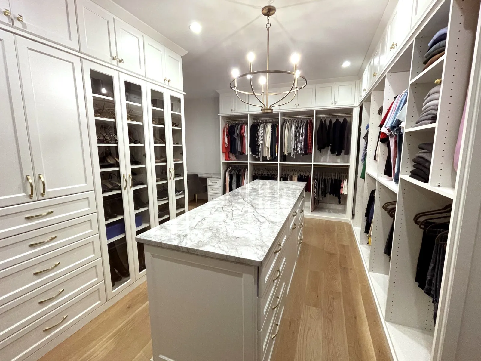 Spacious walk-in closet with white cabinets, marble island, wooden floors, and hanging clothes under chandelier lighting.