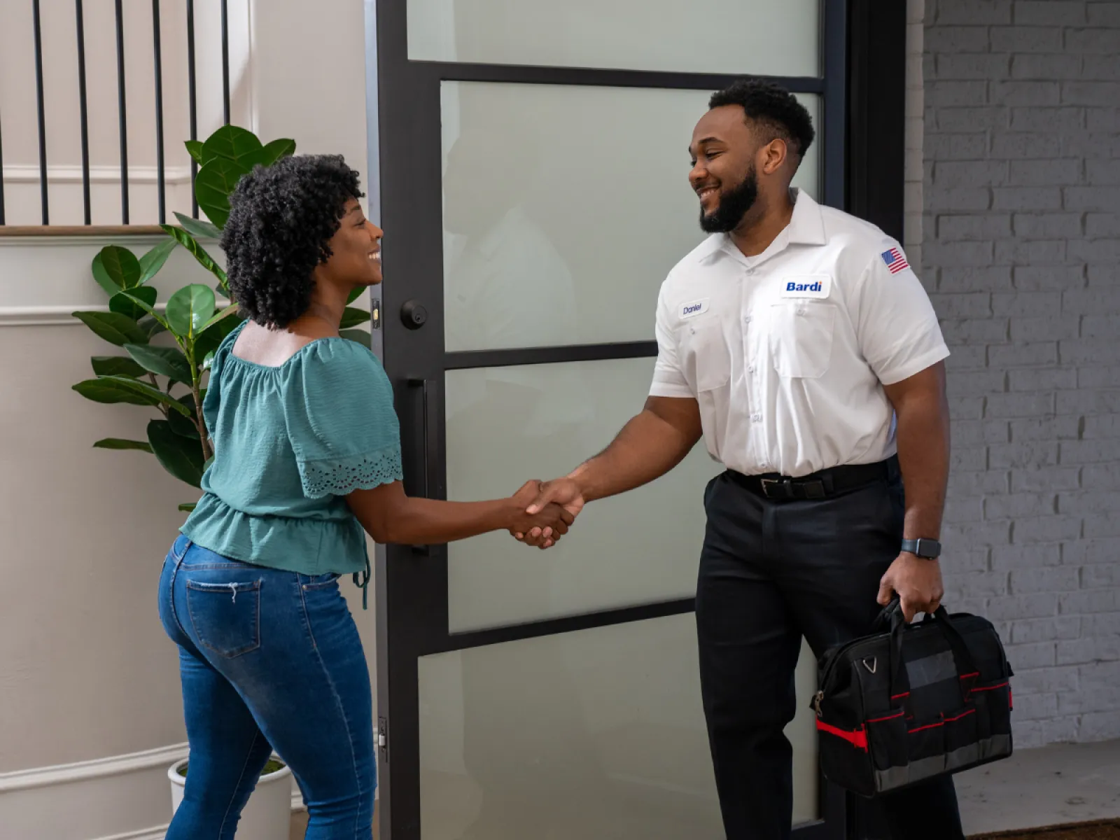Technician in uniform shaking hands with woman at door carrying a black tool bag in a modern home.