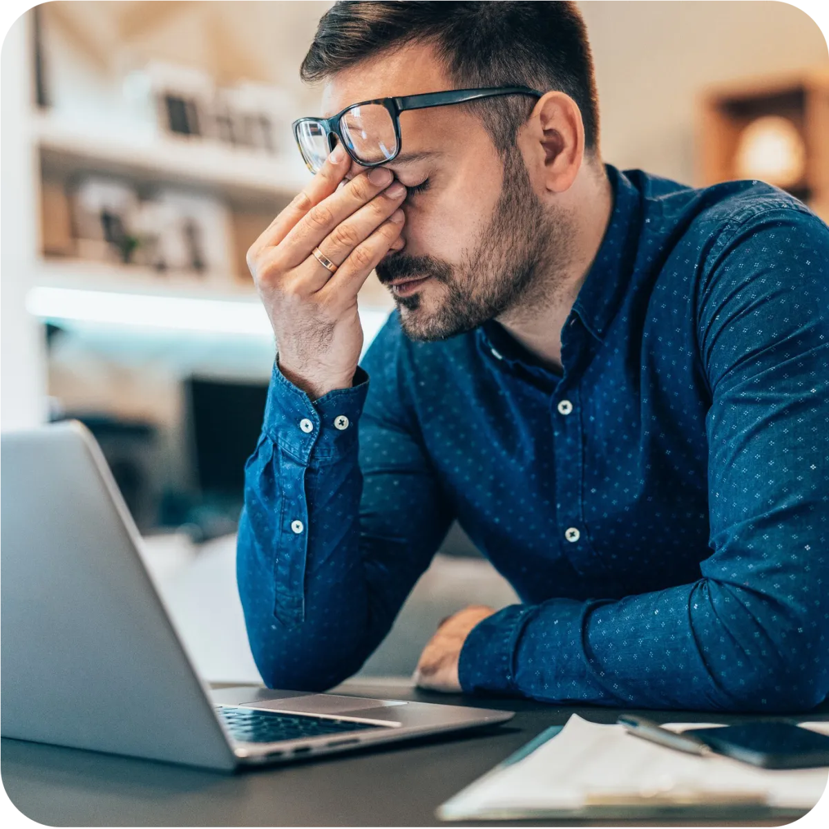 Stressed man in blue shirt rubbing eyes while sitting at desk with laptop and work documents.