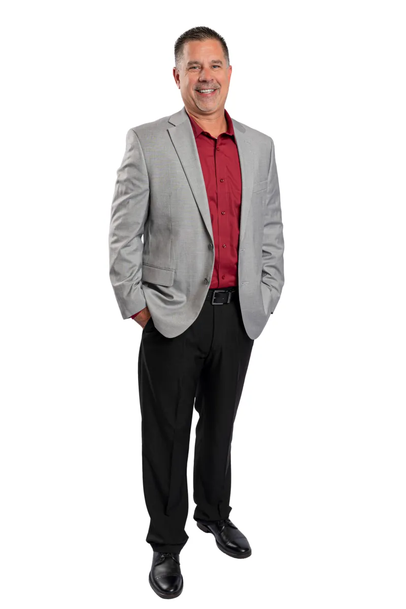 Smiling man in gray blazer, maroon shirt, black pants, and black shoes standing with hands in pockets on white background