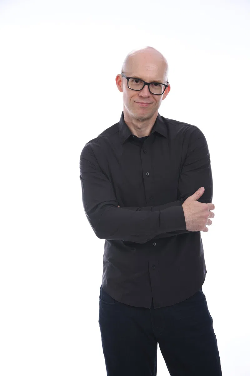 Bald man wearing glasses and black shirt with arms crossed against white background.
