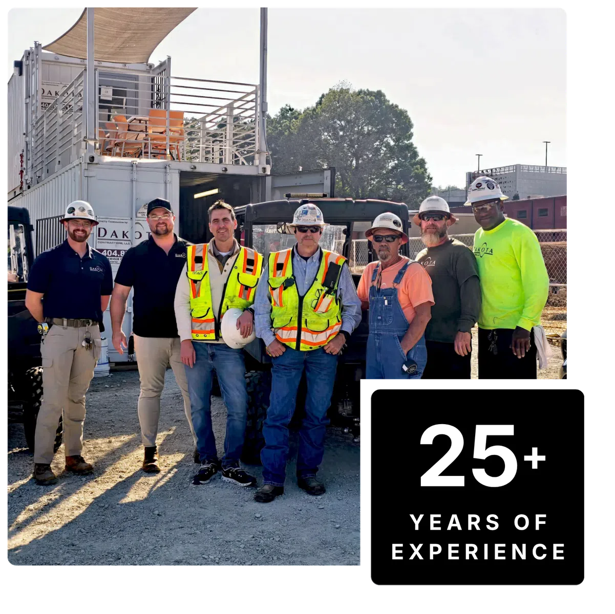 Group of construction workers wearing safety gear standing outdoors on a job site with equipment behind them.