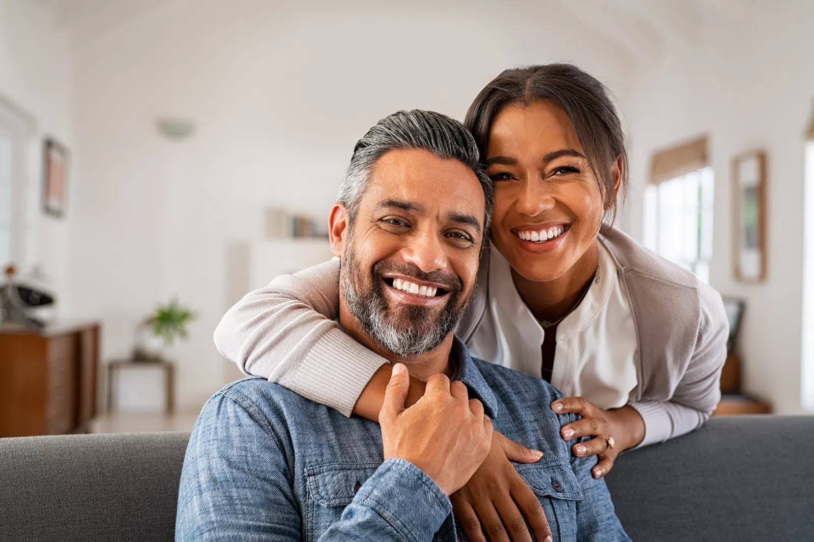 Happy couple smiling and hugging on a couch in a bright living room with cozy decor and natural light.