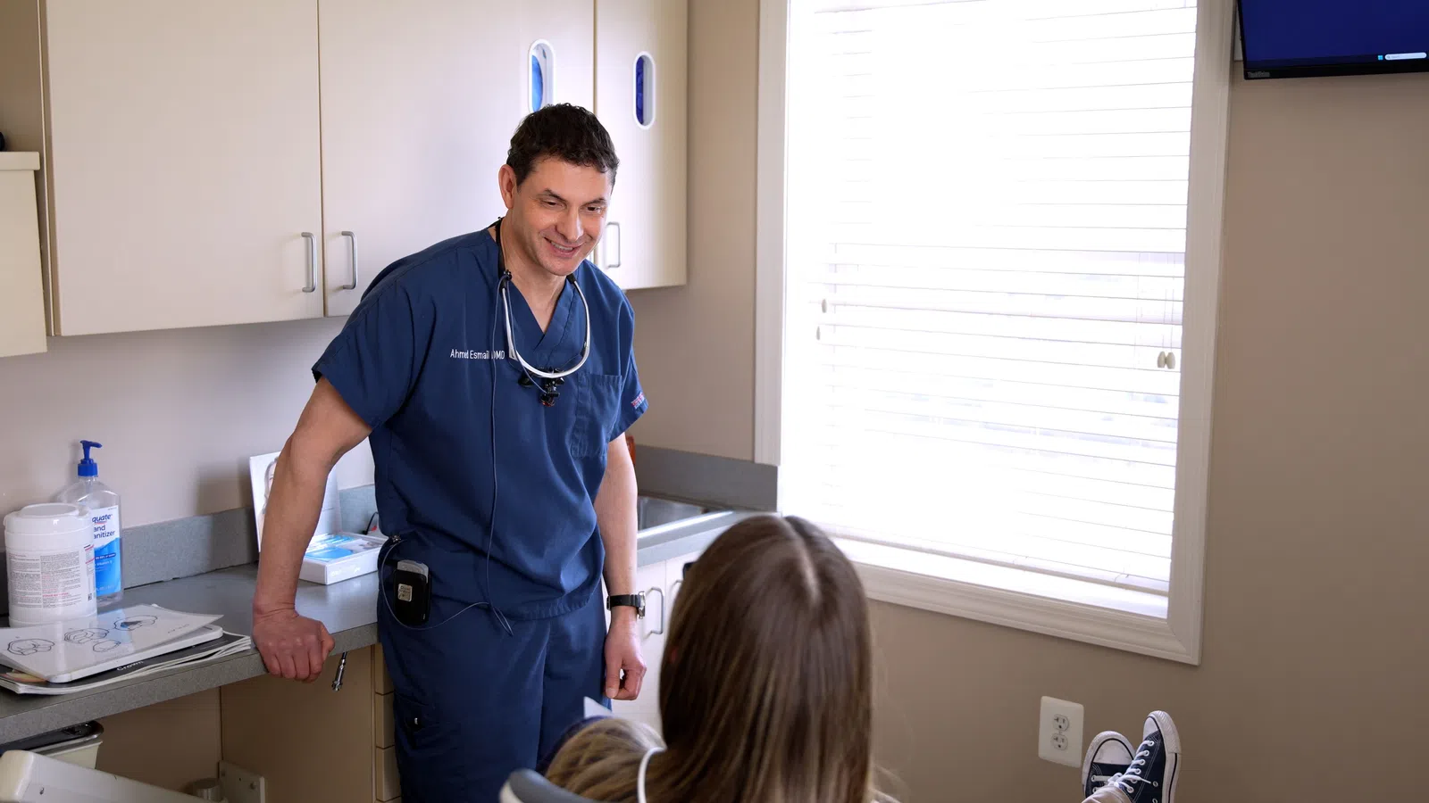 Dentist in blue scrubs consulting with patient in a bright dental office with medical tools visible.