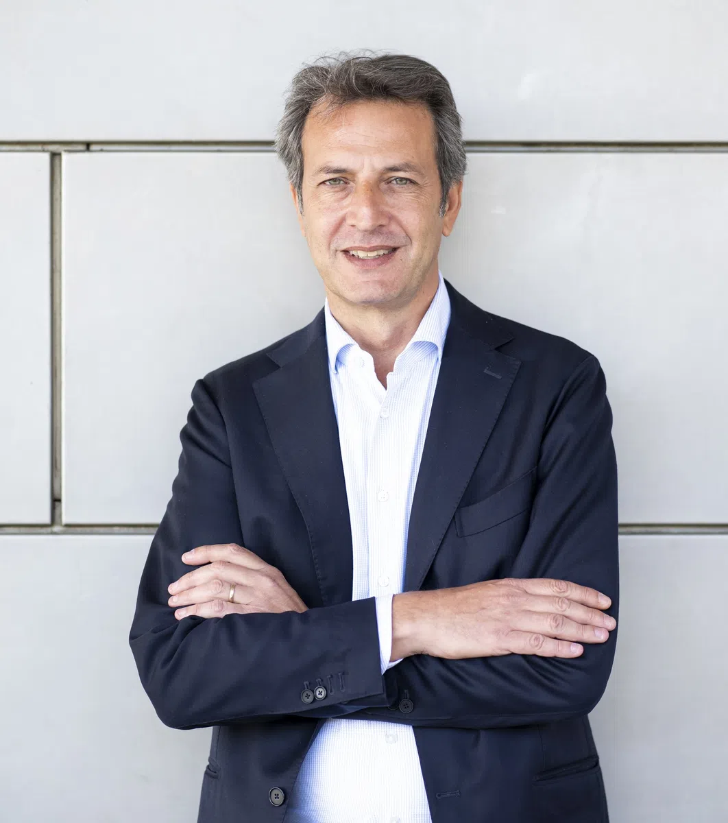 Middle-aged man in a white shirt and black suit jacket standing confidently with arms crossed against a gray wall.