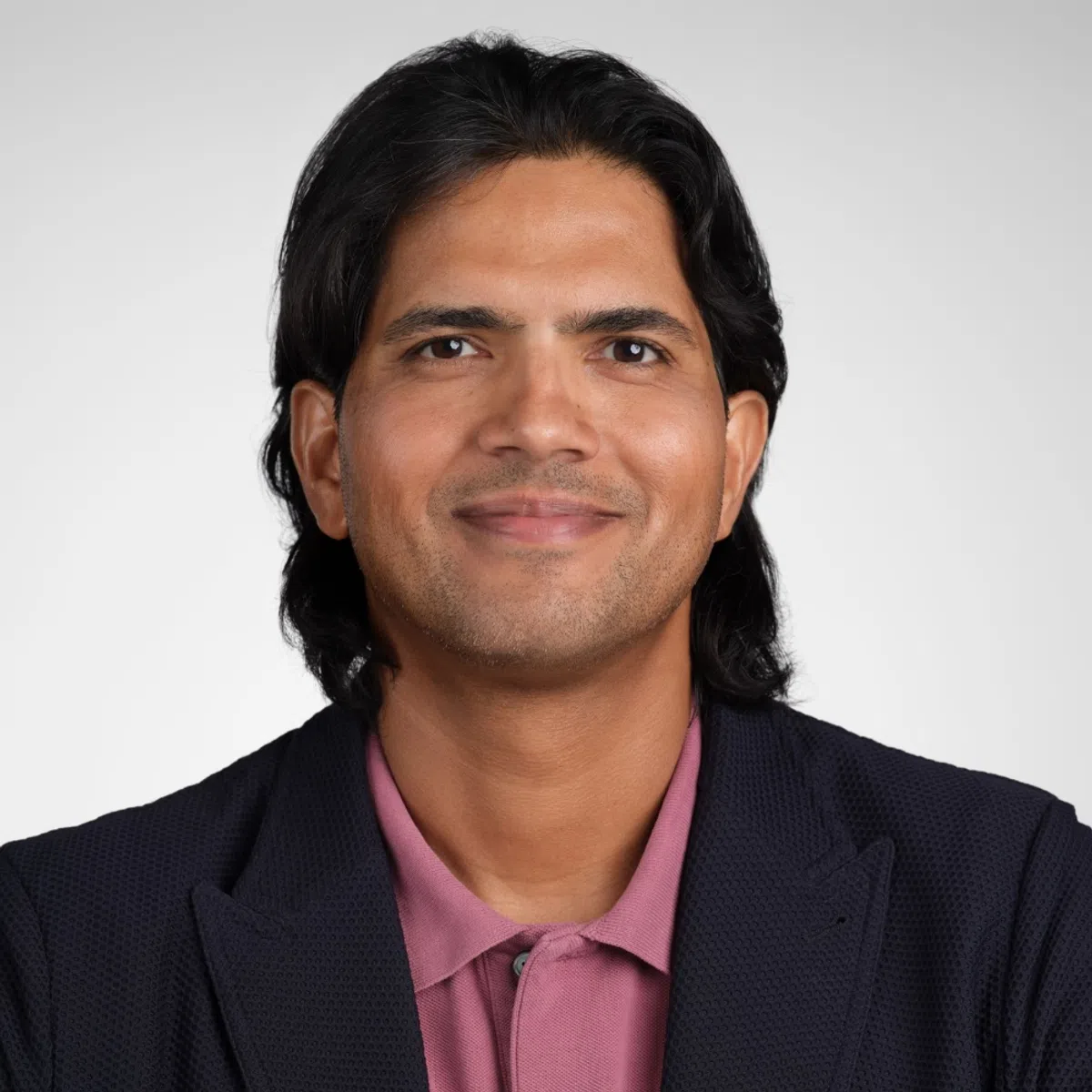 Smiling man with medium-length black hair wearing a pink shirt and dark blazer on a plain gray background