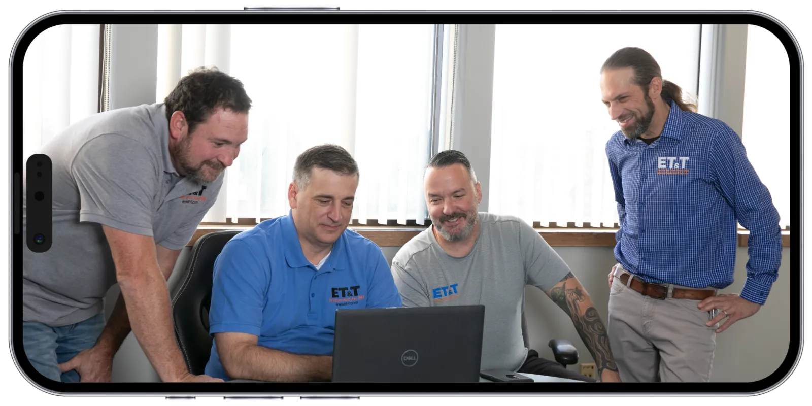 Four men in casual work attire gather around a laptop in a bright office with vertical blinds.