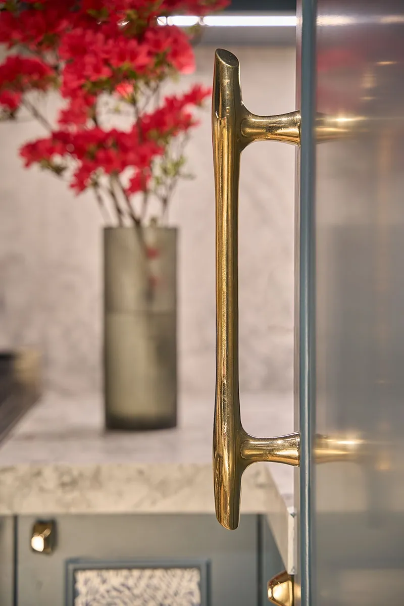 Close-up of a shiny brass door handle on a muted blue door with red flowers blurred in the background