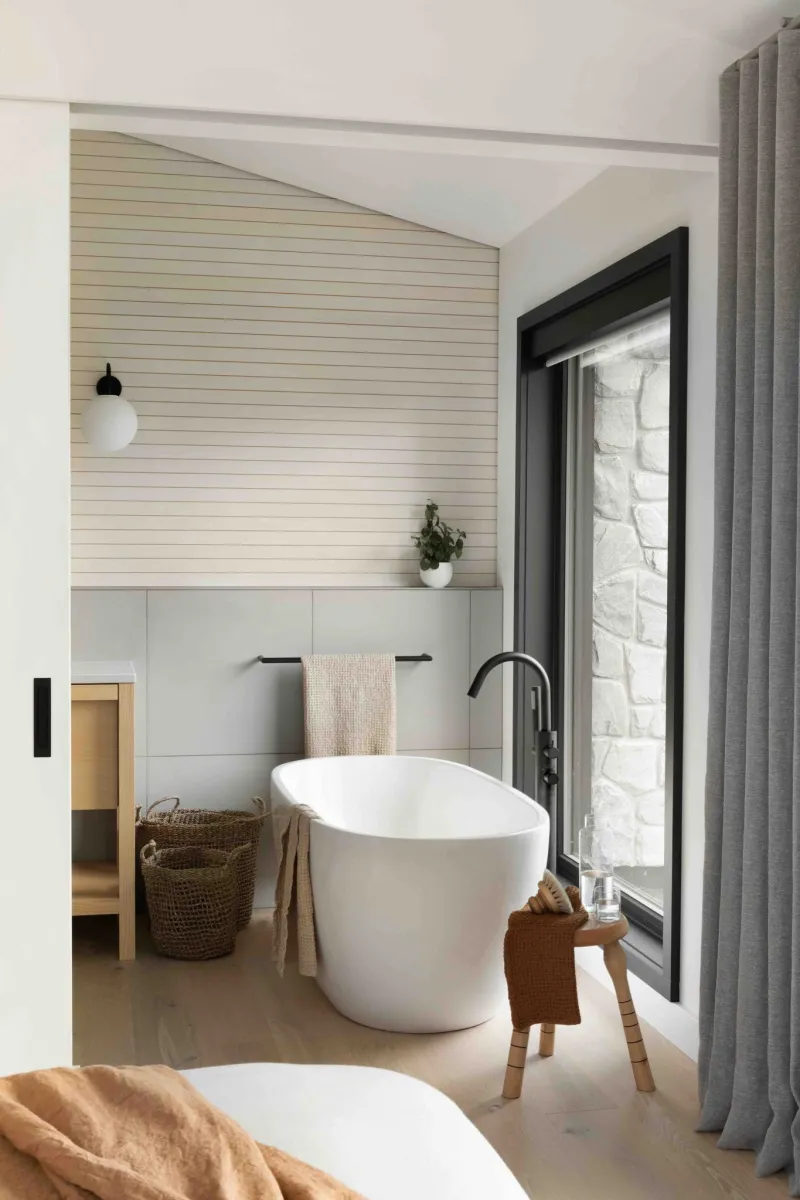 Modern minimalist bathroom with freestanding tub, wooden stool, large window, and neutral decor elements.