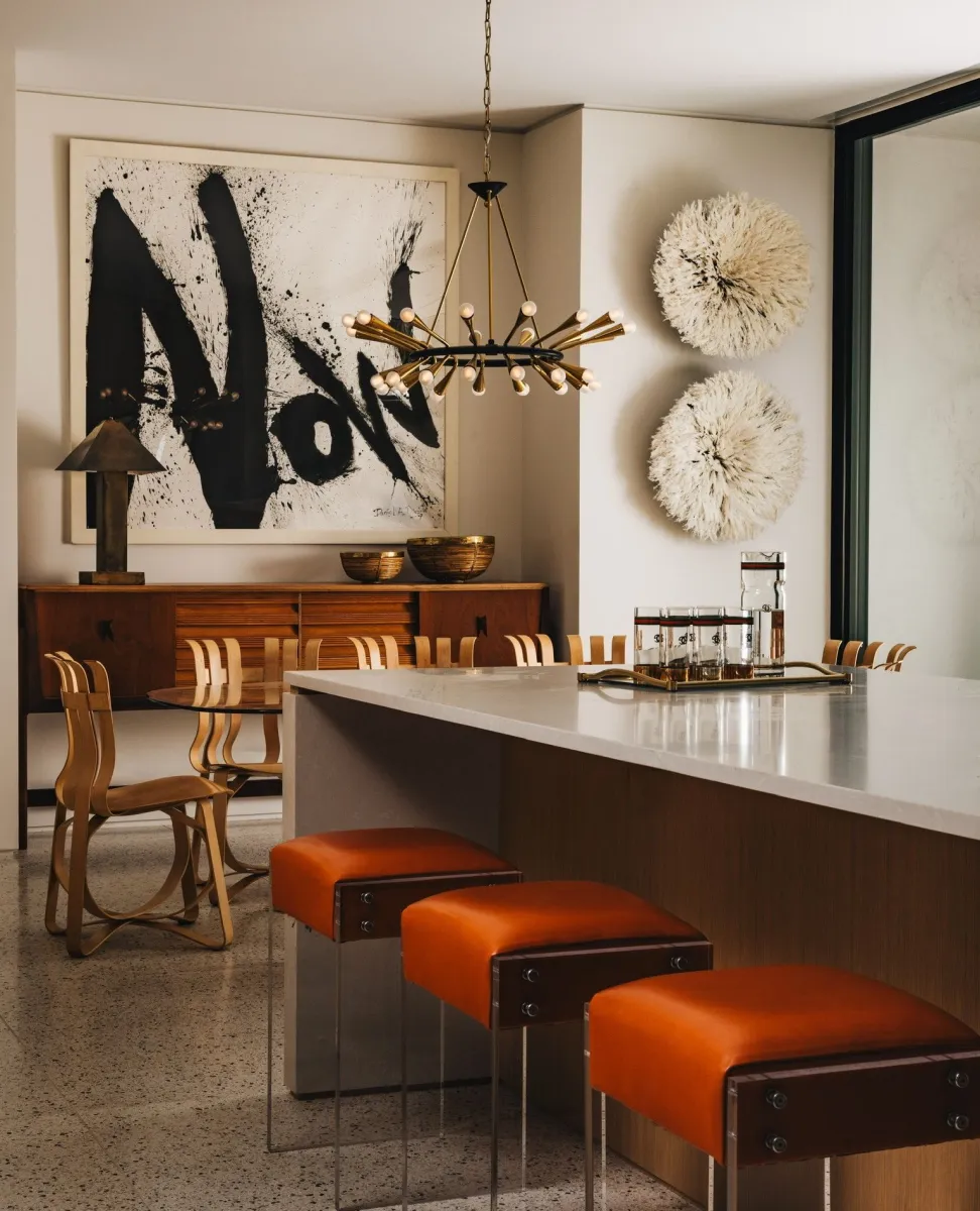 Modern dining area with orange cushioned stools, wooden chairs, abstract black and white art, and a statement chandelier.