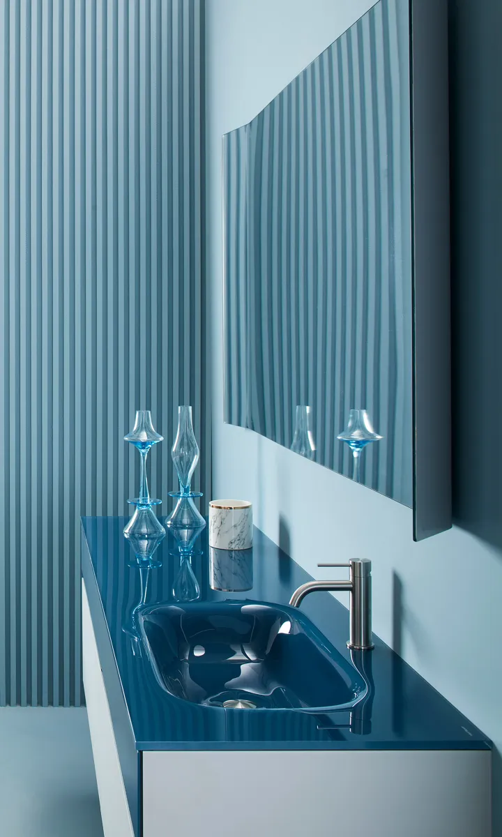Modern bathroom sink with glossy blue countertop, silver faucet, decorative glass vases, and vertical striped wall panel.