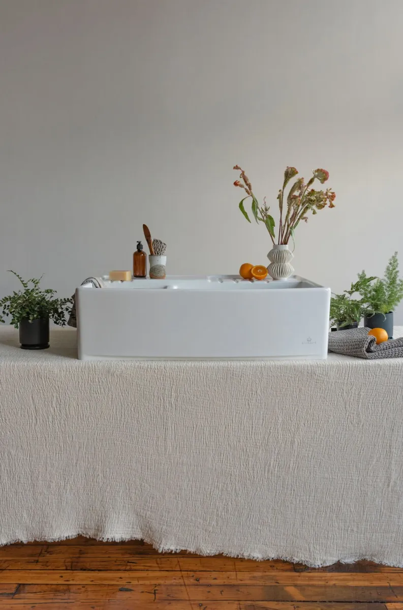 Minimalist white bathtub on textured beige cloth with plants, dried flowers, and oranges in a serene setting.