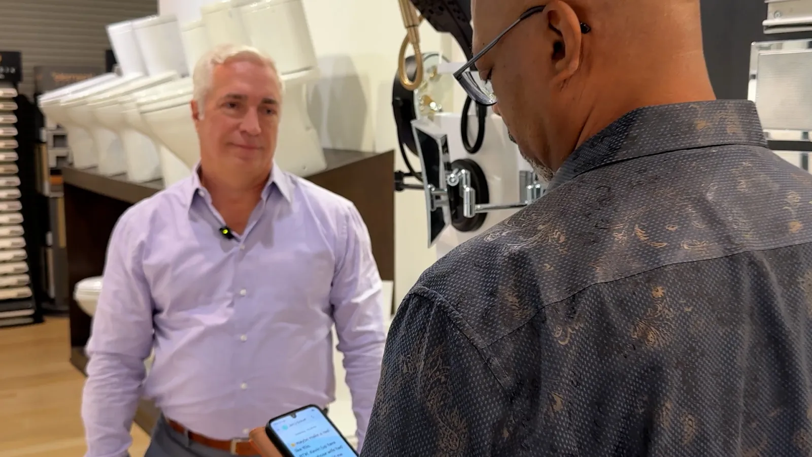 Two men in a showroom discussing products, one holding a smartphone, surrounded by toilet displays.