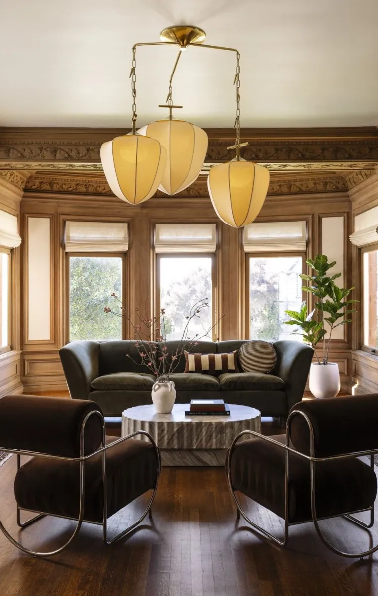 Elegant living room with velvet sofa, modern chairs, sculpted wood paneling, large windows, and unique pendant lights