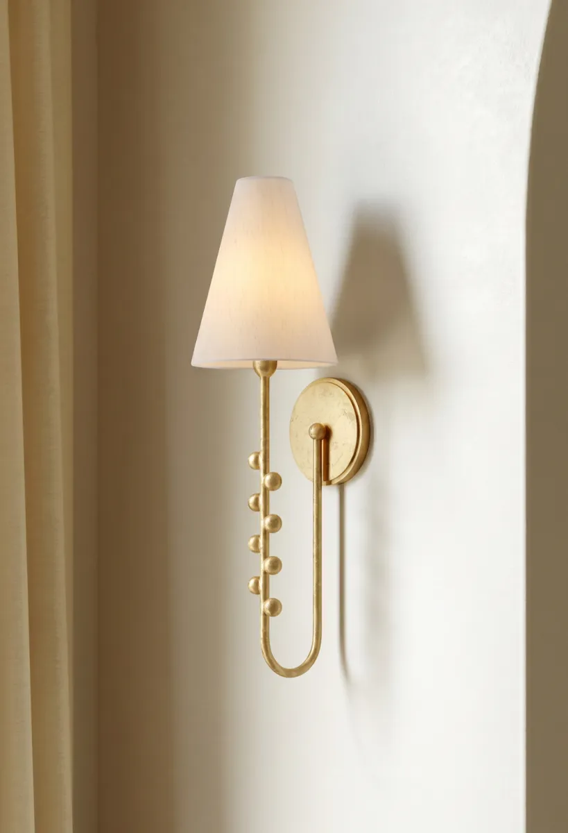 Elegant gold wall sconce with white lampshade mounted on beige textured wall beside curtain and archway