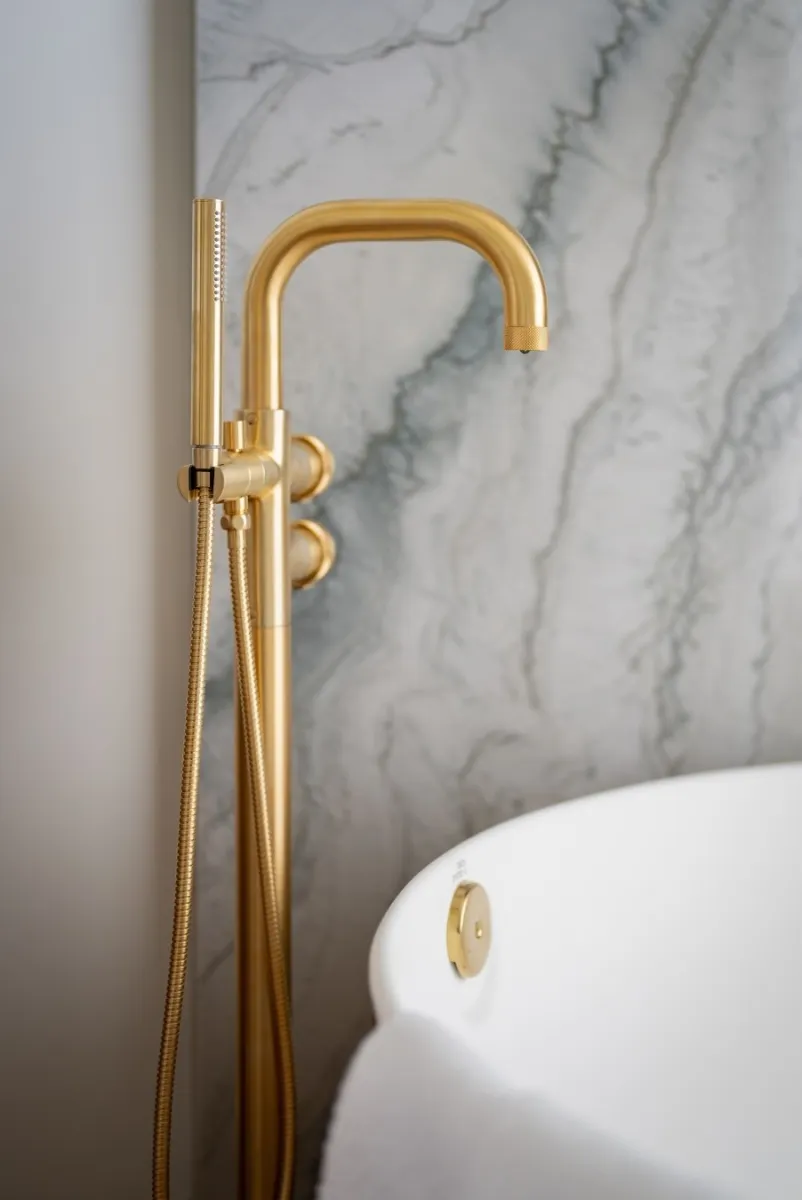 Modern gold bathtub faucet with handheld shower against a gray marble wall and white freestanding tub.