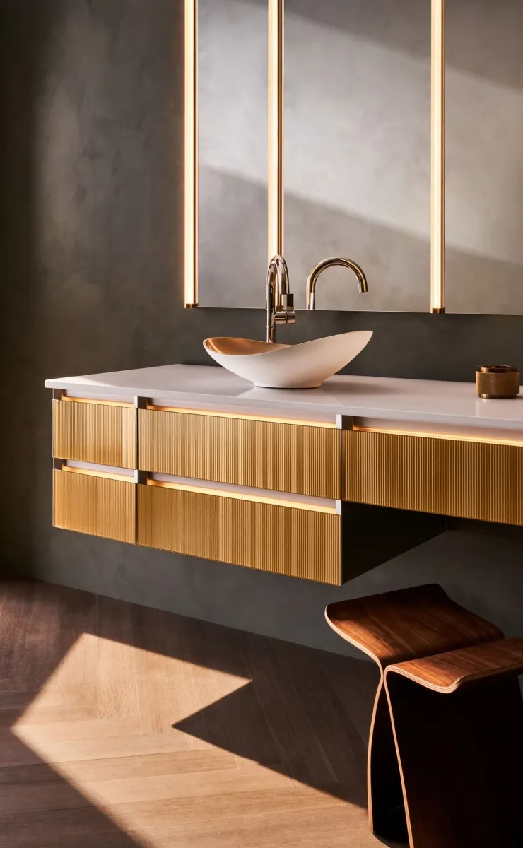 Modern bathroom vanity with gold textured drawers, white vessel sink, lit mirror, and wooden stool on herringbone floor.