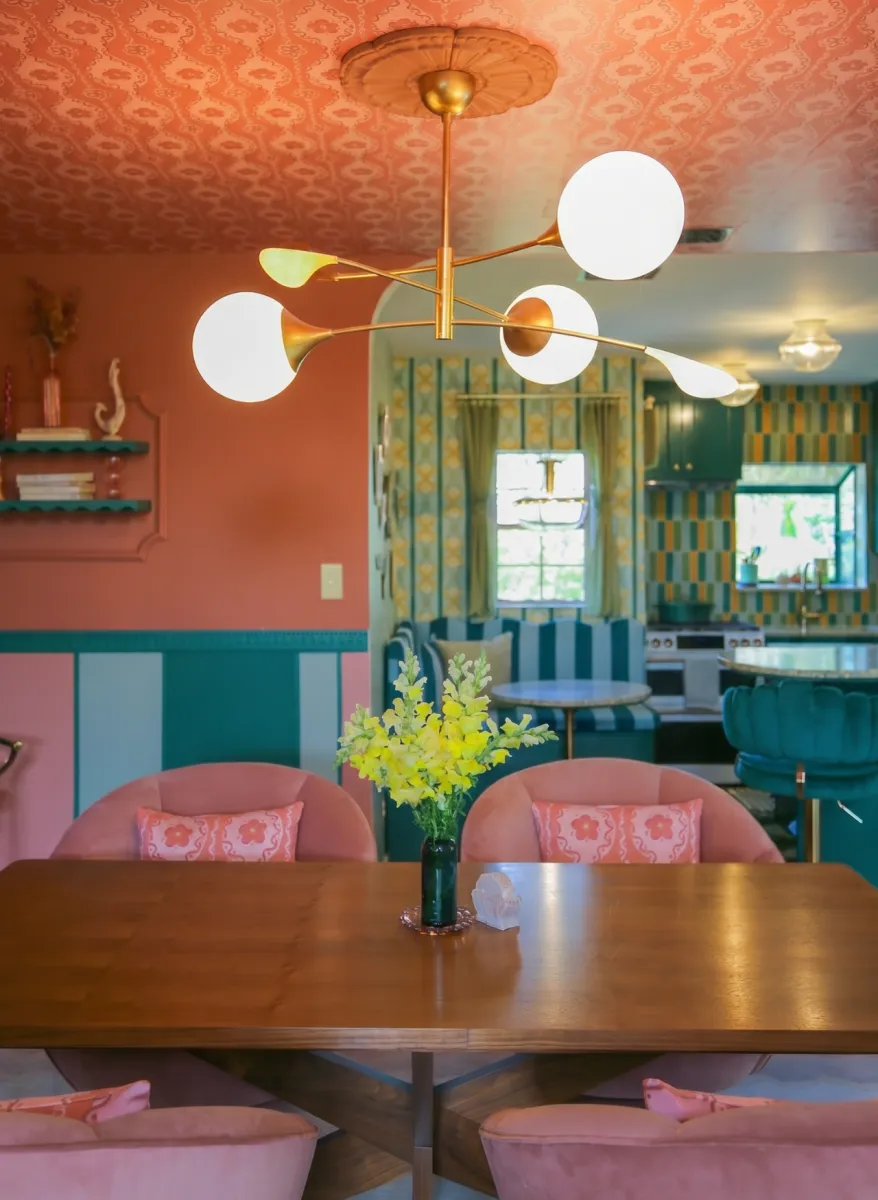 Colorful dining room with pink chairs, wooden table, yellow flowers, modern chandelier, and patterned ceiling.
