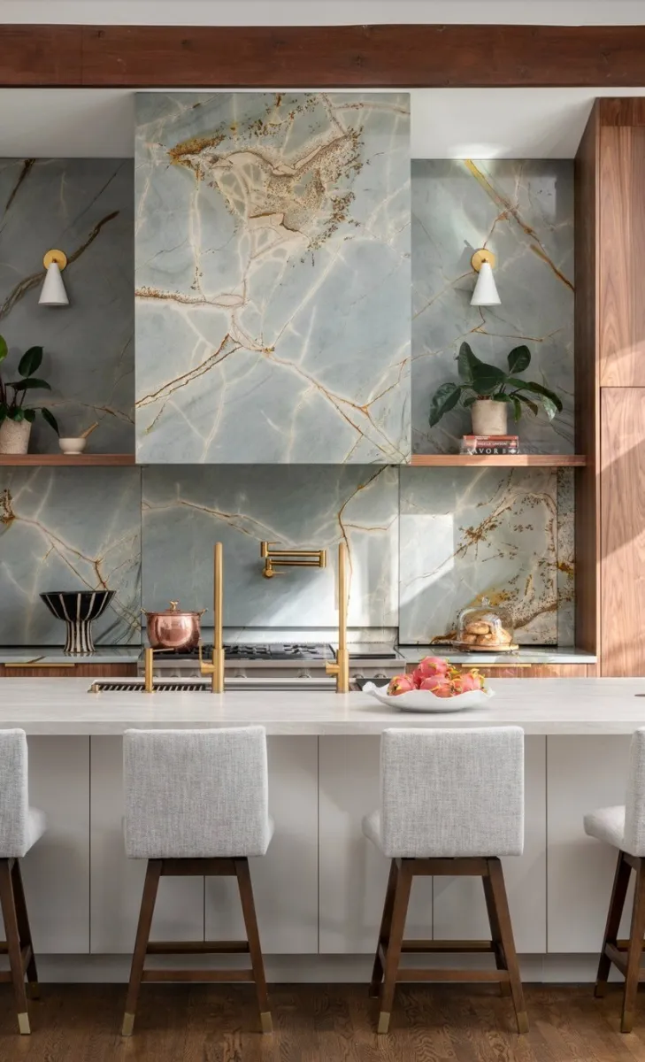 Modern kitchen with marble backsplash, wooden cabinetry, white island, four upholstered stools, and gold fixtures.