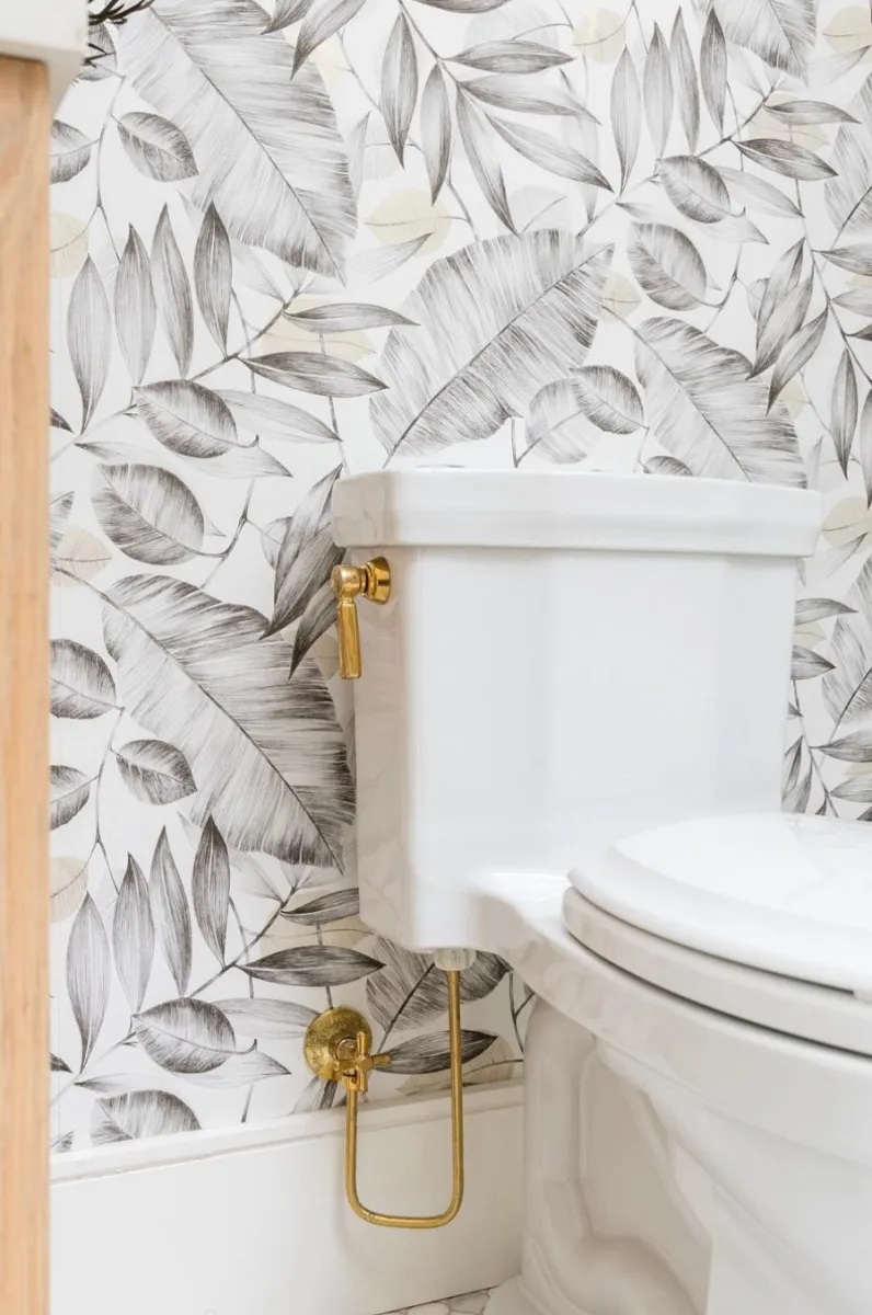 White toilet with gold flush handle in bathroom with gray leaf-patterned wallpaper and wooden cabinet.