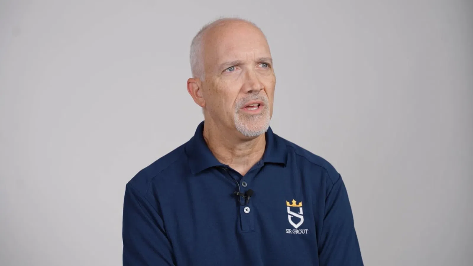 Male franchisee in a navy polo shirt with Sir Grout logo speaking against a gray background.