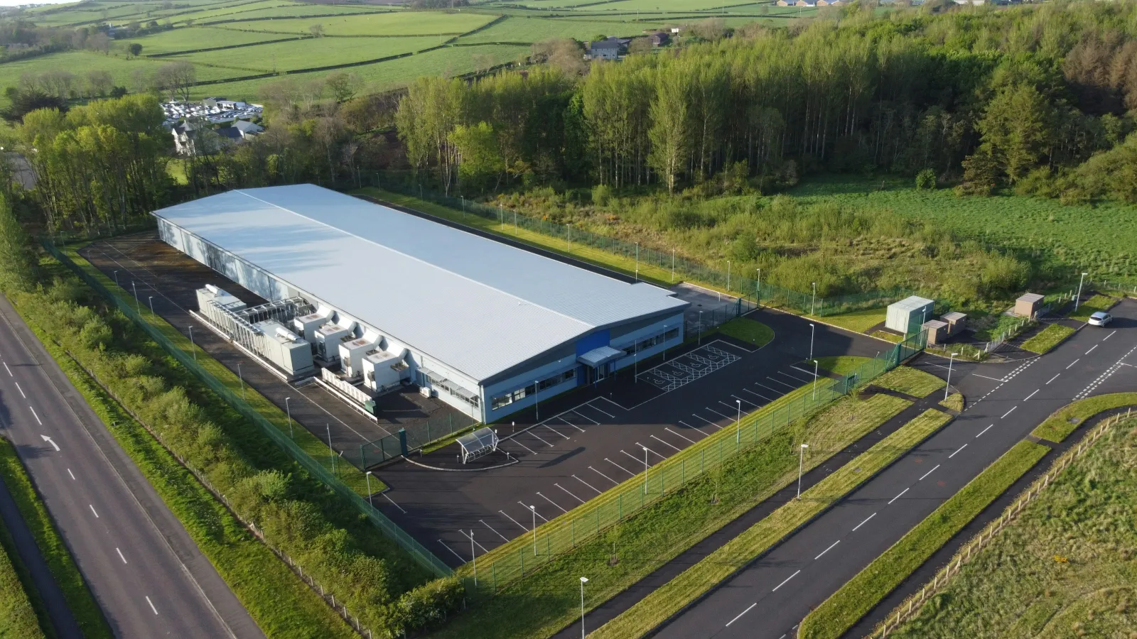 Aerial view of a large industrial warehouse surrounded by greenery and adjacent roads on a sunny day.