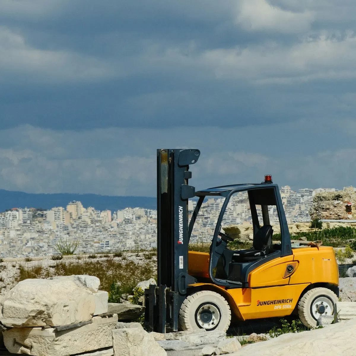 Yellow Jungheinrich forklift parked among large stone blocks with cityscape and cloudy sky in background