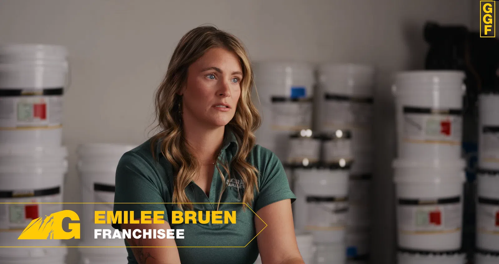 Woman in green shirt named Emilee Bruen franchisee sitting with paint buckets behind her in a warehouse setting.