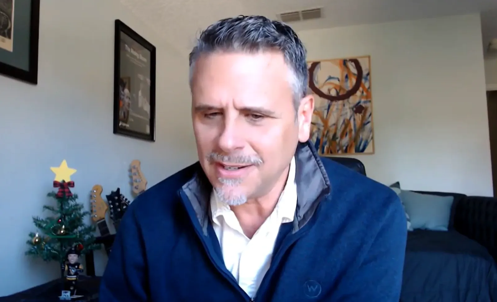 Man with salt-and-pepper hair and goatee speaking indoors with Christmas tree and guitars in background