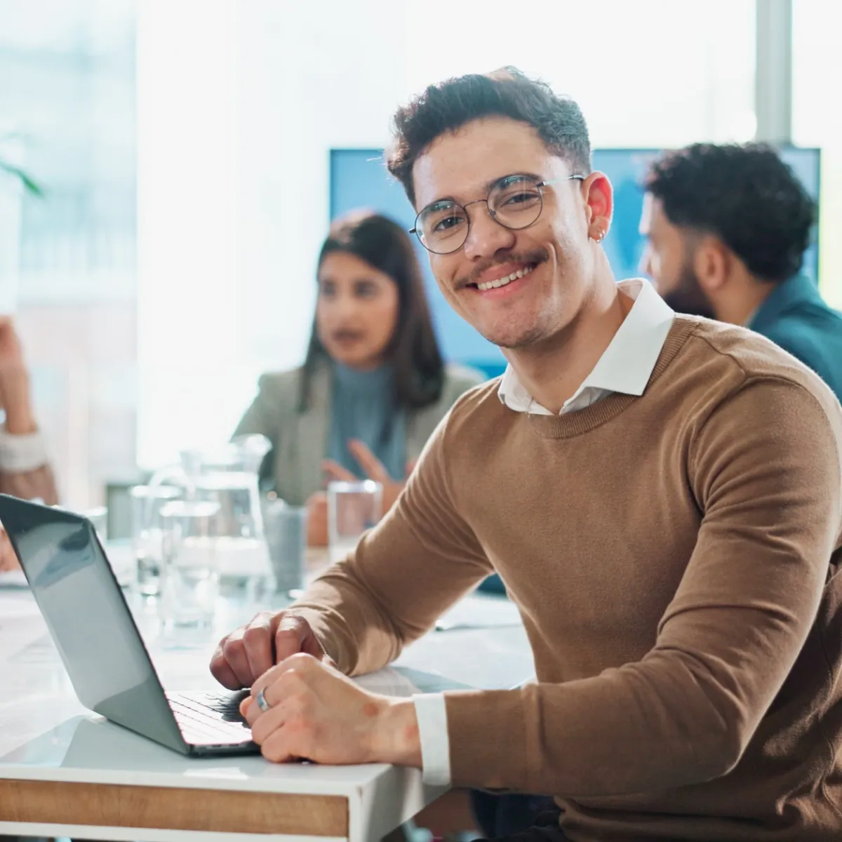 Young professional man smiling at laptop in a modern office meeting with diverse colleagues in the background