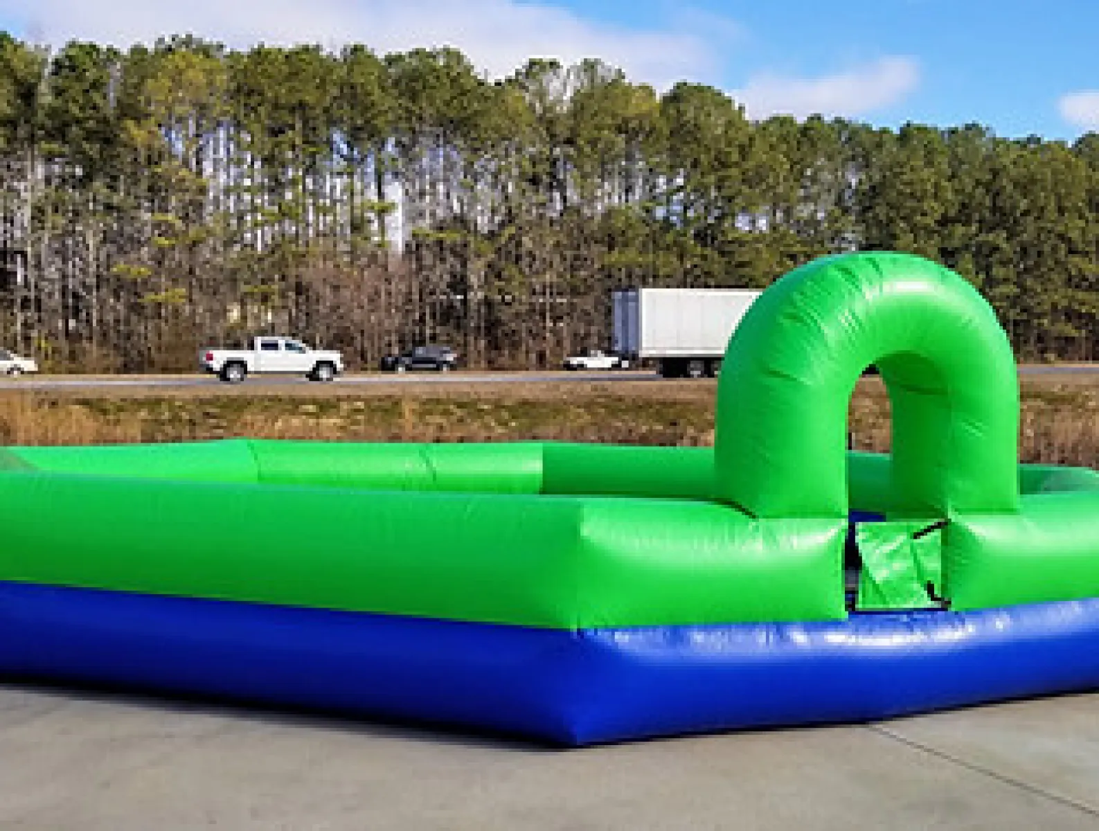 Large green and blue inflatable soccer field set up outdoors on concrete near a highway with trees in background
