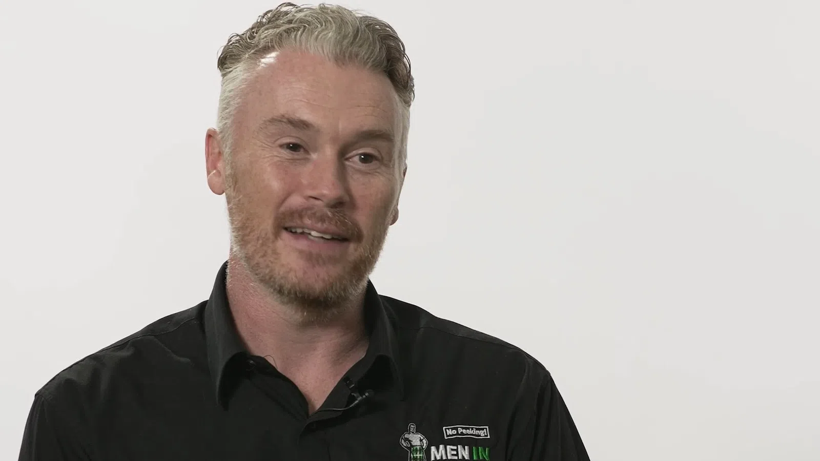 Smiling man with light hair wearing a black collared shirt with a logo on plain light background.