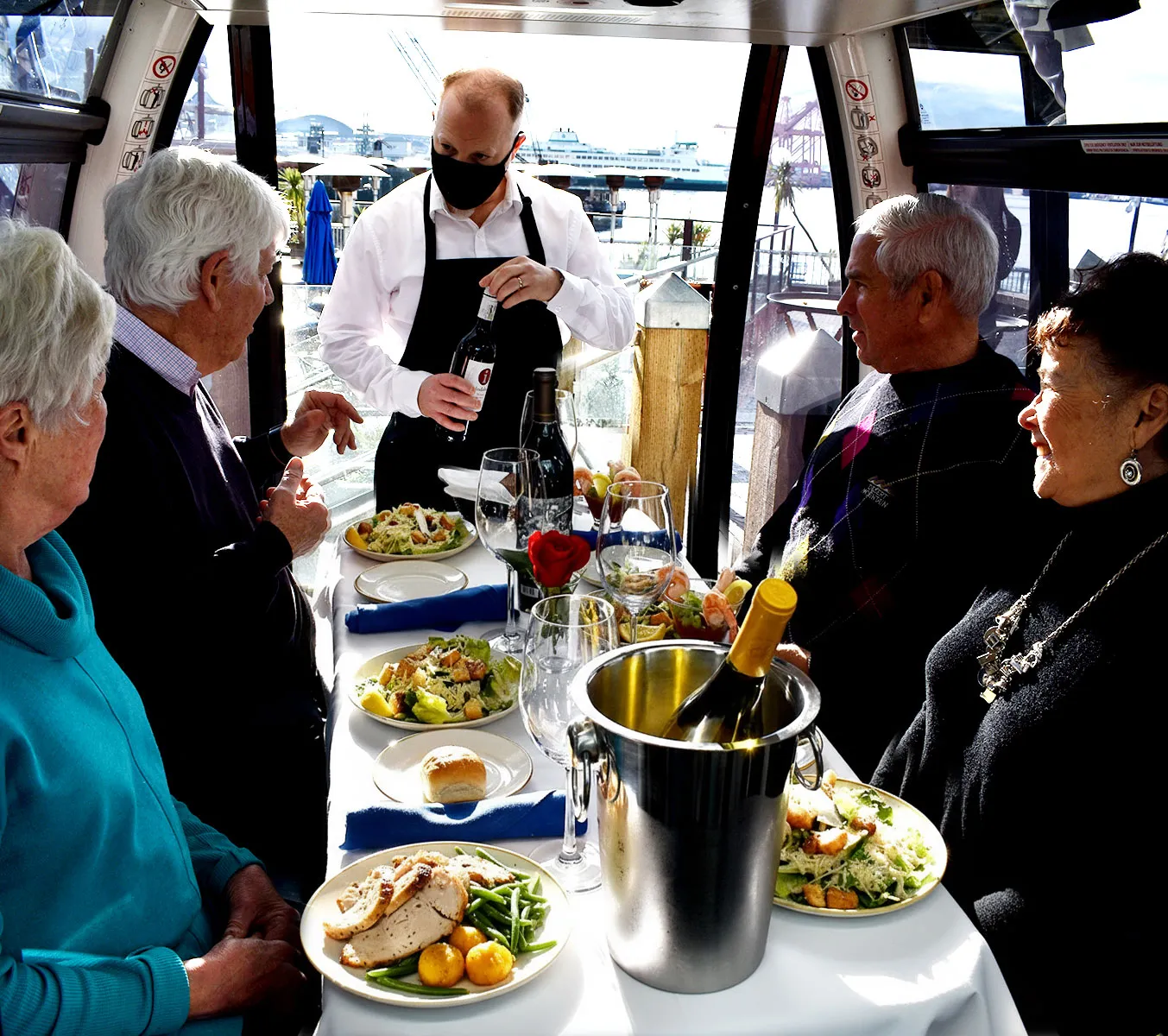 Waiter serving wine to four seniors enjoying a meal with salads, chicken, and wine on a boat with water views.
