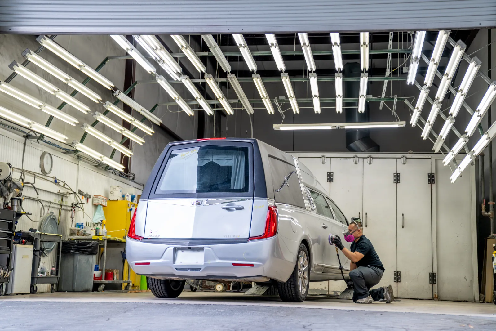Person polishing a silver hearse inside a brightly lit workshop with numerous fluorescent lights overhead.