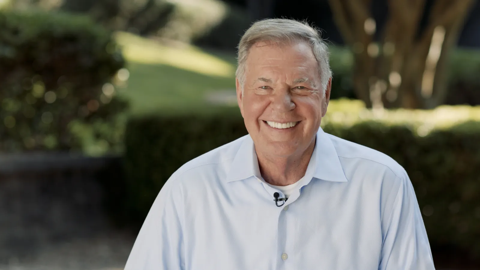 Smiling elderly man wearing a light blue shirt outdoors with blurred greenery background.