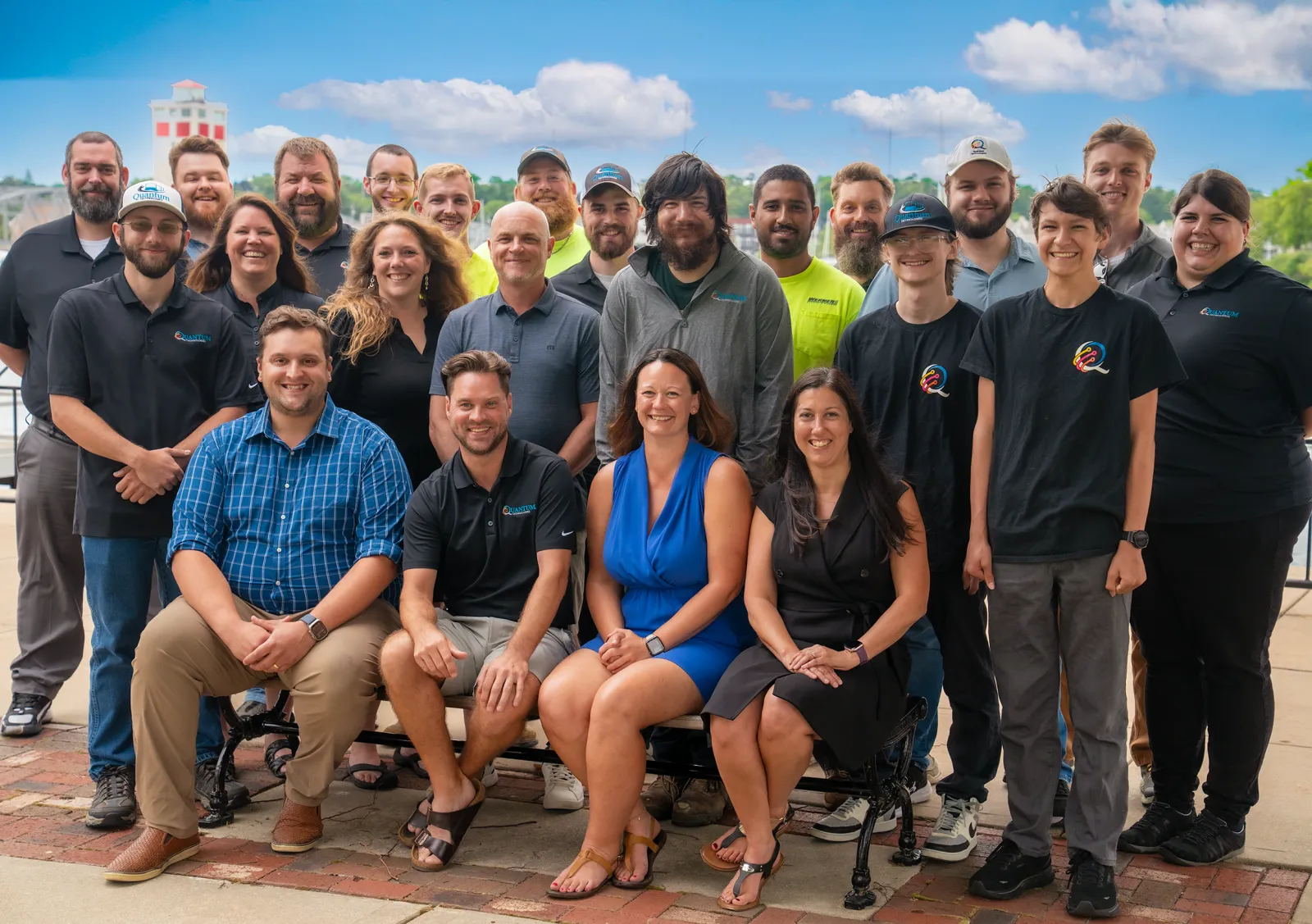Group portrait of diverse smiling adults posing outdoors on a brick patio with a lighthouse and blue sky background.