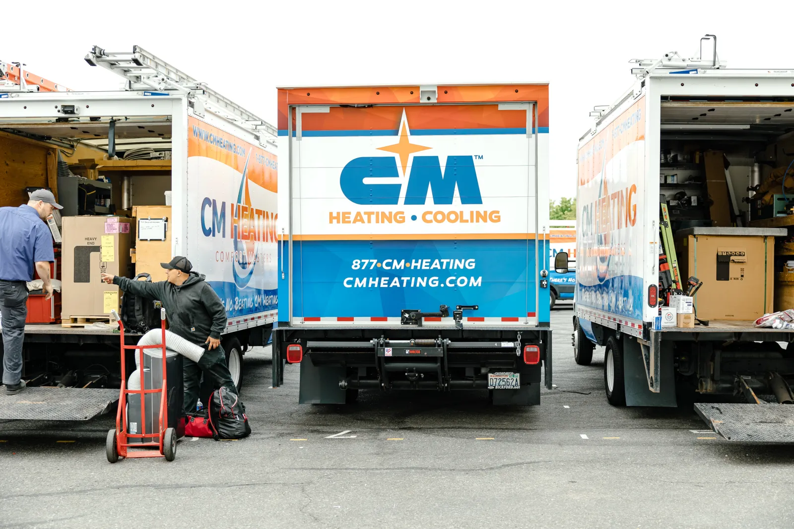 Two workers unloading equipment from CM Heating and Cooling trucks in a parking lot during daytime.