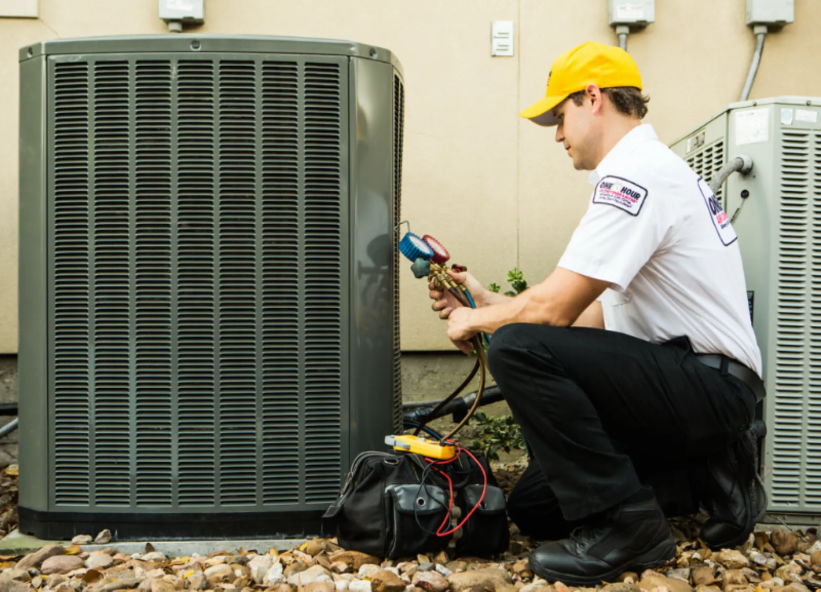 Technician in yellow hat servicing outdoor air conditioning unit with gauges and tools on rocky ground