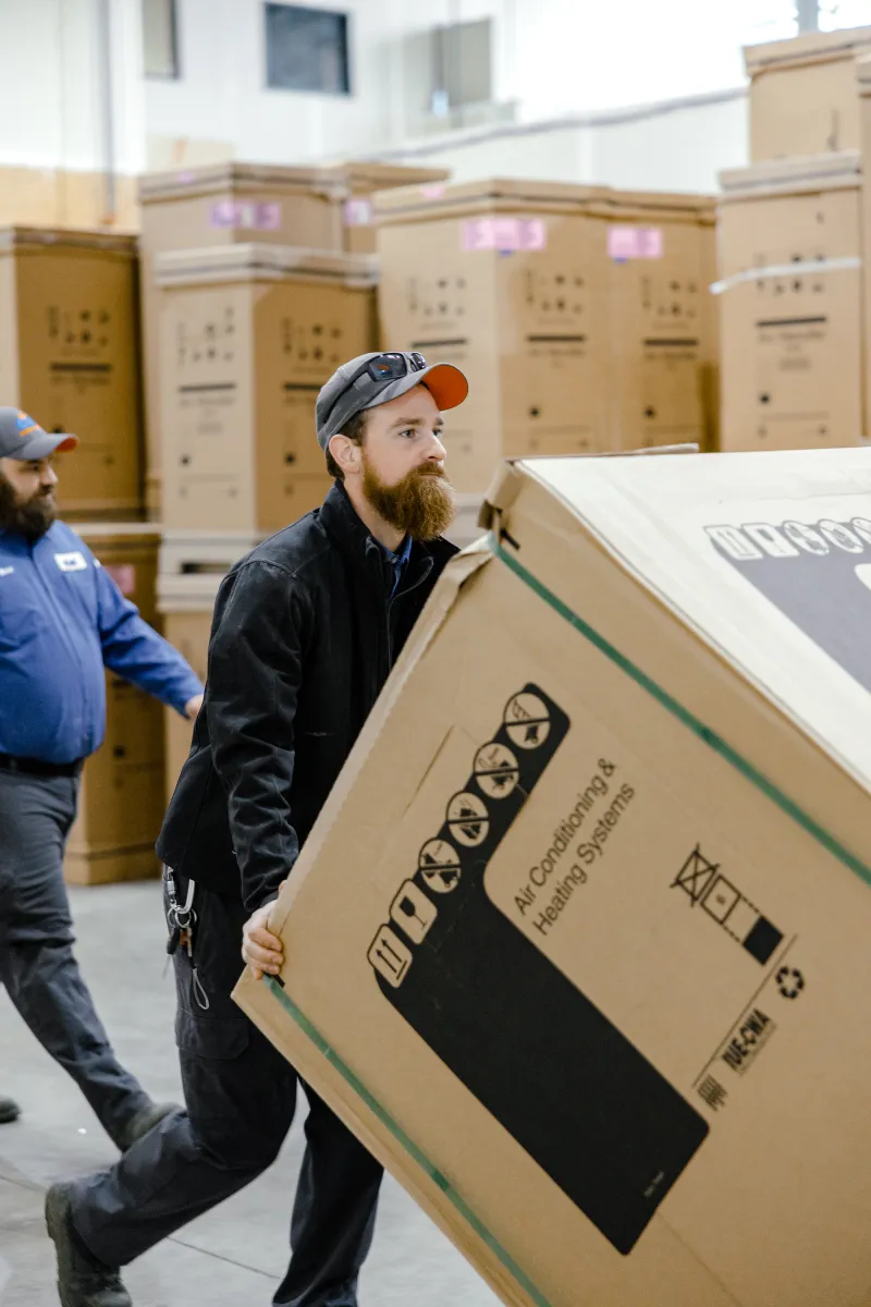 Worker carrying a large air conditioning and heating system box in a warehouse with stacked boxes in background.